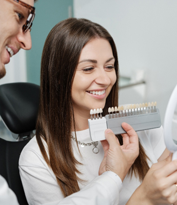 a patient looking in a handheld mirror while a dentist holds a shade chart in front of her teeth